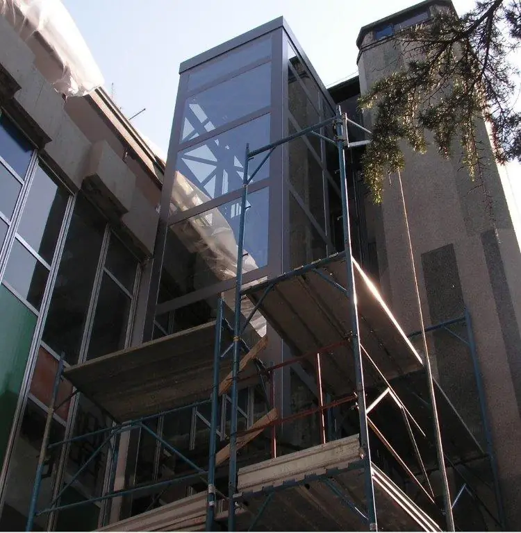 Torre ascensore esterna in acciaio e vetro affiancata a un condominio